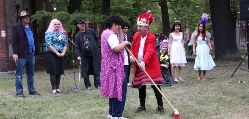 Ein König und eine Putzfrau stehen auf einer Wiese. Im Hintergrund weitere verkleidete Personen.