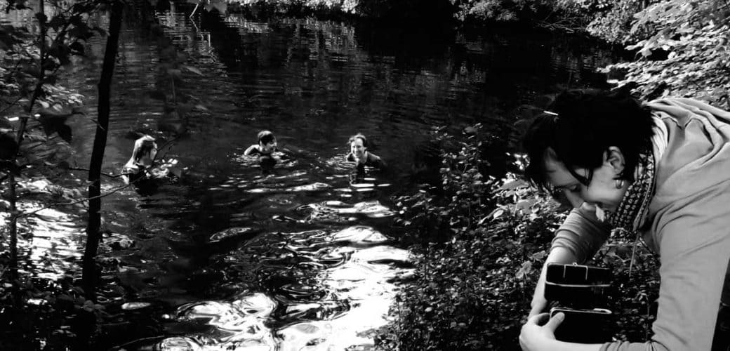 Drei fröhliche Menschen in einem See. Eine Frau mit Kamera im Vordergrund. Schwarz-weiß-Bild.
