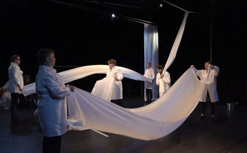 Szene aus dem Theaterstück. Menschen in weißen Kitteln schwenken weiße Tuchbahnen.